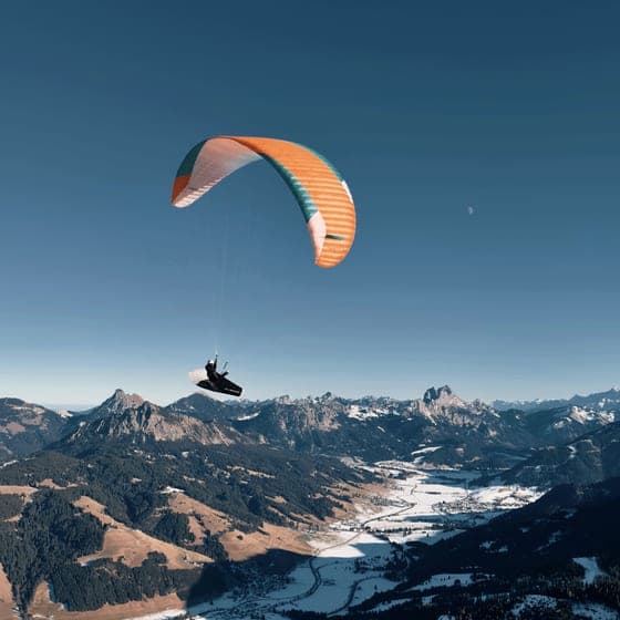 Gleitschirm hoch über einem verschneiten Alpental, darunter gestaffelte Bergketten