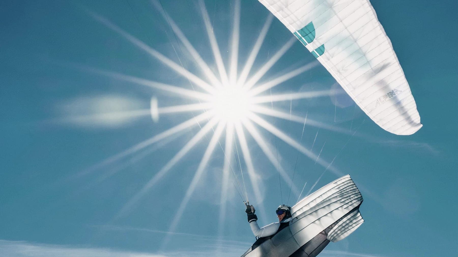 Gleitschirm im Gegenlicht über einer verschneiten Kette, die Sonne strahlenförmig dahinter